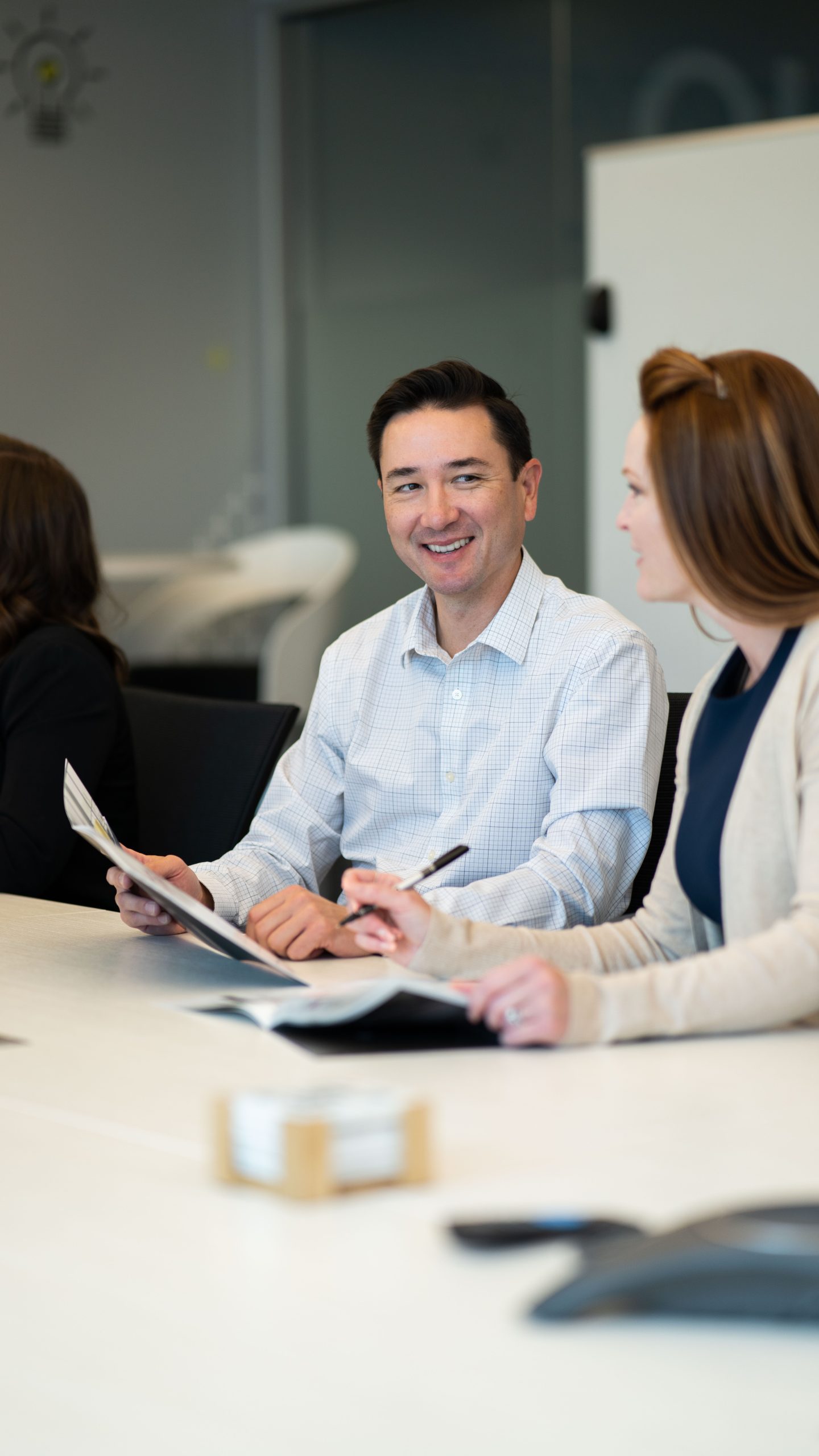 Professional meeting in a modern office setting, featuring a smiling man in a checkered shirt engaged in conversation with a woman, while another person is partially visible in the background. The table is equipped with documents and a pen, indicating a collaborative work environment.