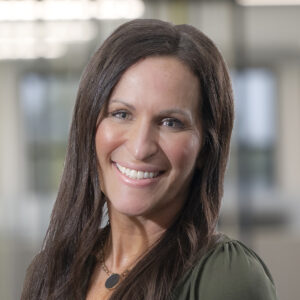 Smiling woman with long brown hair and a green blouse, standing in a well-lit modern office environment.