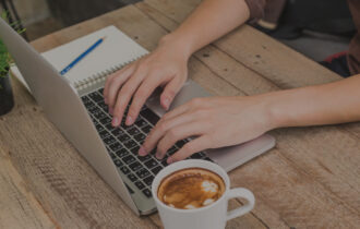 Hands typing on a laptop placed on a light wooden table with a cup of coffee in a white mug and a notepad and pen next to the laptop