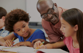 Male teacher leans over students who are writing in notebooks to help them with what they are working on