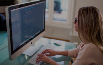 Woman in a pink sweater sits at a teal blue desk with her hands on her keyboard as she codes on the desktop in front of her.