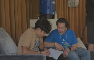 Man sitting next to young boy while he reads from notebook.