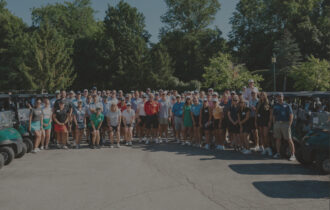 A large group of people in golf clothes gather for a photo with golf carts to their side.