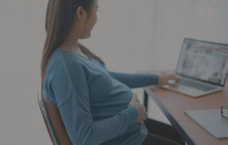 A pregnant woman sits at her desk looking at her laptop with her hand over her stomach.